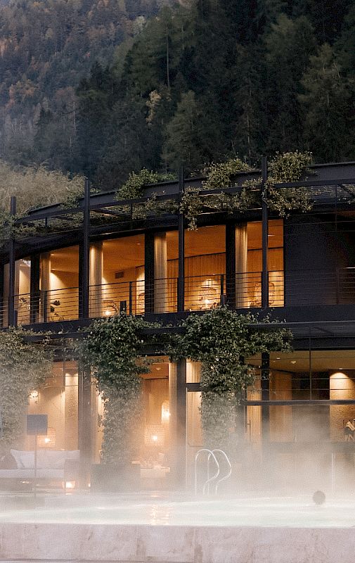 Edificio moderno con facciata in vetro, circondato da boschi, con interni illuminati al tramonto.