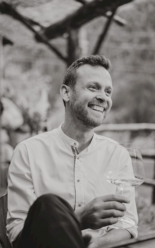 A man in a shirt smiling, holding a wine glass. Background shows a wooden fence.
