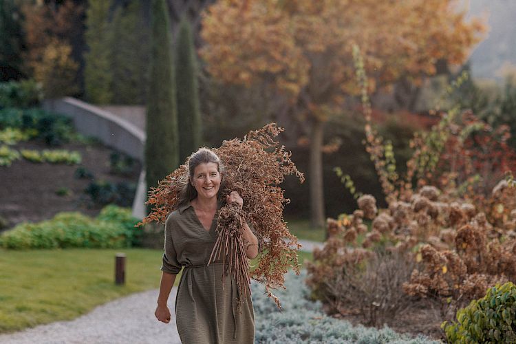 Frau trägt im herbstlichen Garten einen Haufen trockener Zweige, umgeben von buntem Laub.