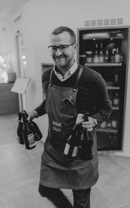 Ein lächelnder Mann in einer Schürze trägt mehrere Weinflaschen in einem stilvollen Raum.