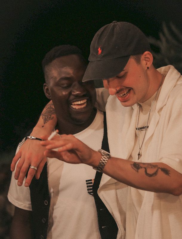 Due uomini, sorridendo, si abbracciano in un'atmosfera rilassata e informale.