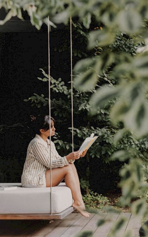 Frau liest ein Buch auf einer Schaukel im Garten, umgeben von grünen Pflanzen.