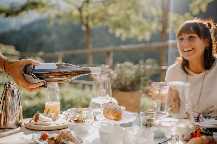 Ein Tisch im Freien mit Essen und Getränken, jemand schenkt Wein ein, eine lächelnde Frau im Hintergrund.