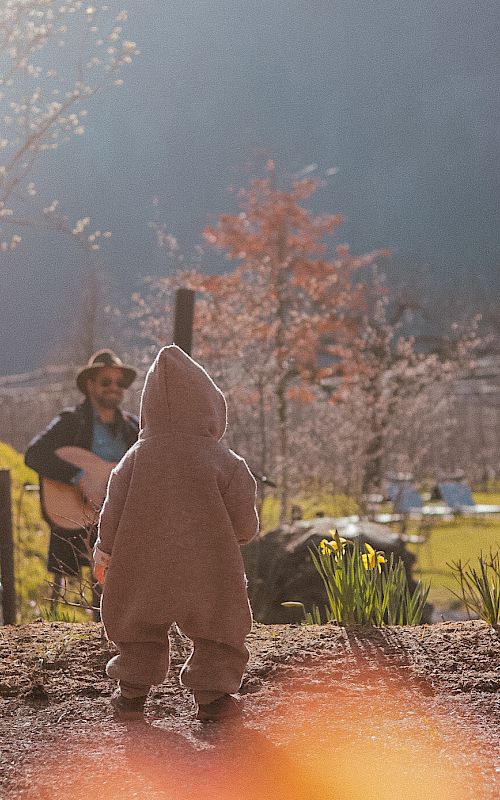 Kleinkind draußen in dickem Anzug blickt auf Musiker im Hintergrund, Frühlingstag mit Blumen.